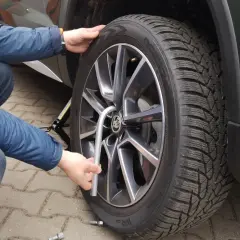 Osoba w niebieskiej kurtce dokręca śrubę koła samochodu kluczem.