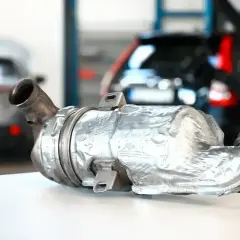 Używany filtr DPF na białym stole, w tle warsztat samochodowy z autami.