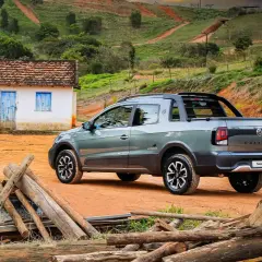 Szary Volkswagen Saveiro Extreme na tle wiejskiego krajobrazu z małym domkiem i stertą drewna.