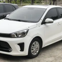 Biały sedan Kia Pegas z przyciemnianymi szybami zaparkowany obok czarnego SUV-a.
