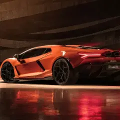 Pomarańczowy Lamborghini Revuelto z czarnymi felgami, stojący w ciemnym, betonowym garażu, z czerwonymi światłami odbijającymi się od podłogi.