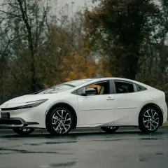 Biały Toyota Prius C na mokrej drodze, otoczony jesiennymi drzewami.