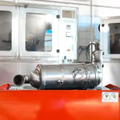 Metalowy katalizator samochodowy leży na pomarańczowej skrzyni na tle urządzeń przemysłowych.