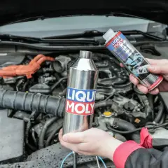 Ręce trzymające preparaty Liqui Moly do czyszczenia katalizatora na tle silnika samochodu.