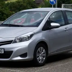 Srebrny Toyota Yaris P13 zaparkowany na kostce brukowej, z widocznym czerwonym samochodem w tle.