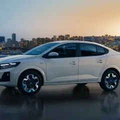 Jasnoszary Dacia Supernova jedzie po mokrej drodze z widokiem na nocne miasto w tle.