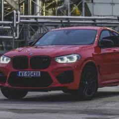 Czerwone BMW X4 z czarnymi felgami i grillem, zaparkowane przed przemysłowym budynkiem.