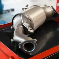 Metalowy element układu wydechowego samochodu, prawdopodobnie filtr DPF, leżący na wózku warsztatowym.