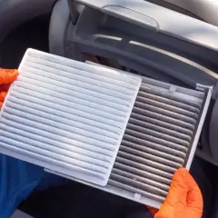 Ręce w pomarańczowych rękawiczkach trzymają nowy, biały filtr kabinowy obok starego, brudnego filtra w samochodzie.
