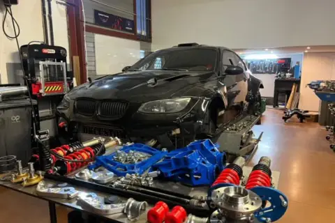 Warsztat z czarnym BMW E92, częściami zawieszenia i narzędziami. Lata produkcji tego modelu to 2006-2013.