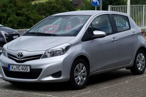 Srebrny Toyota Yaris P13 zaparkowany na kostce brukowej, z widocznym czerwonym samochodem w tle.