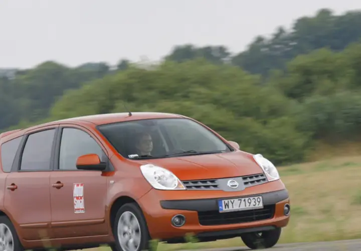 Pomarańczowy Nissan Note E12 z kobietą za kierownicą jedzie po drodze wśród zieleni.