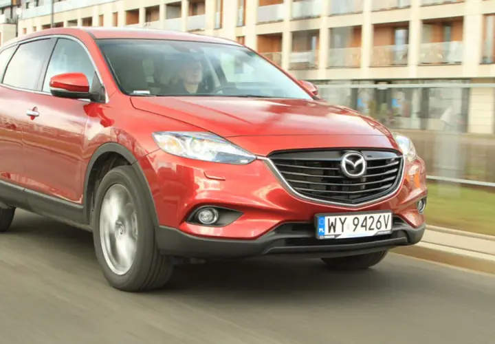 Czerwona Mazda CX-9 w ruchu miejskim, z widocznym kierowcą za kierownicą.