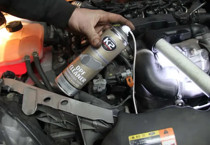 Ręka trzymająca puszkę preparatu do czyszczenia DPF na tle silnika samochodowego.