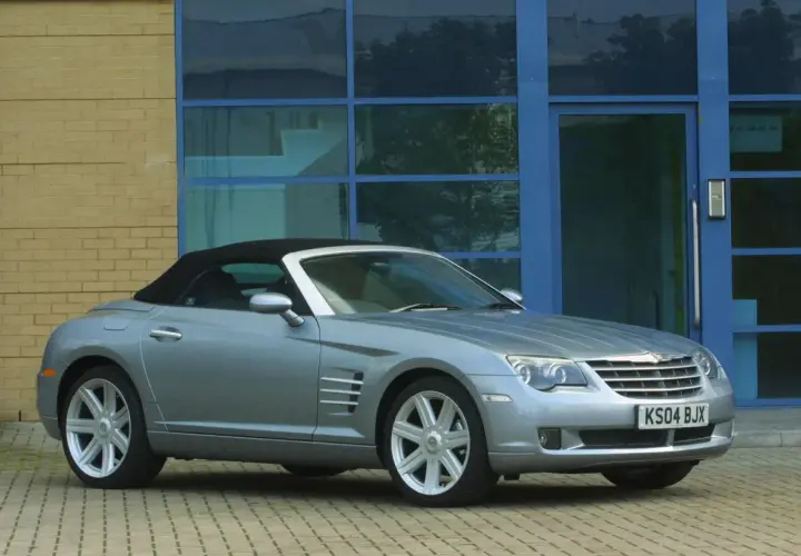 Srebrny Chrysler Crossfire auto z czarnym dachem stoi przed budynkiem z żółtej cegły i niebieskimi oknami.