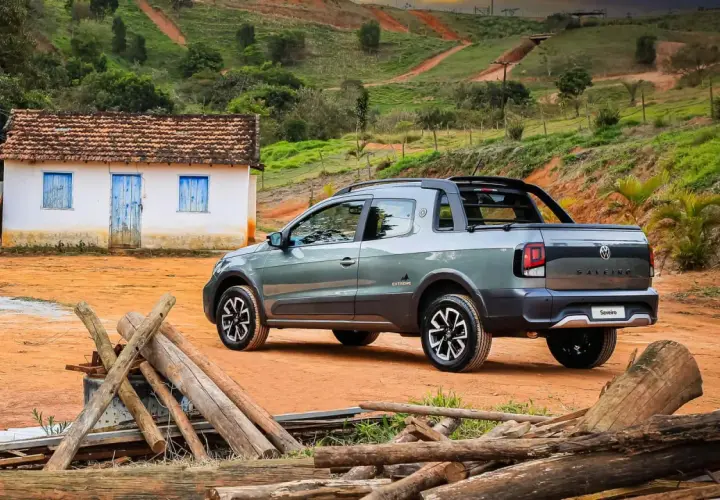 Szary Volkswagen Saveiro Extreme na tle wiejskiego krajobrazu z małym domkiem i stertą drewna.