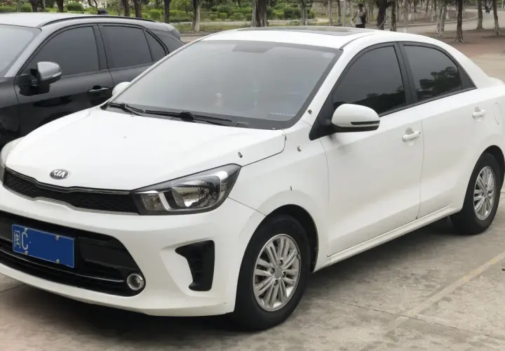 Biały sedan Kia Pegas z przyciemnianymi szybami zaparkowany obok czarnego SUV-a.