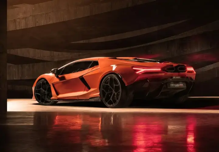 Pomarańczowy Lamborghini Revuelto z czarnymi felgami, stojący w ciemnym, betonowym garażu, z czerwonymi światłami odbijającymi się od podłogi.