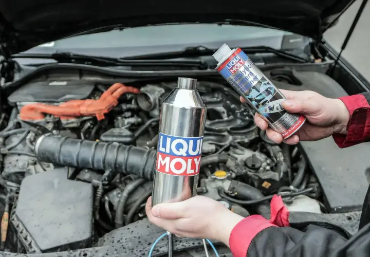 Ręce trzymające preparaty Liqui Moly do czyszczenia katalizatora na tle silnika samochodu.