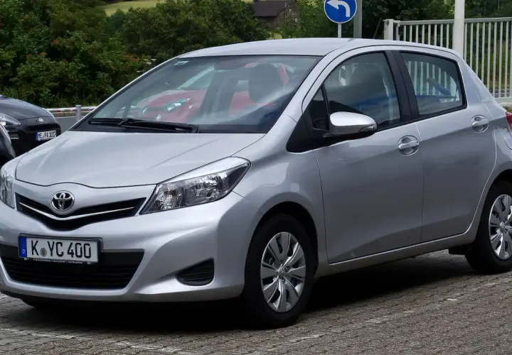 Srebrny Toyota Yaris P13 zaparkowany na kostce brukowej, z widocznym czerwonym samochodem w tle.