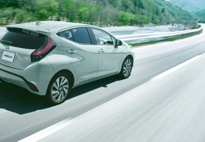 Jasnoszary Toyota Aqua jedzie po krętej drodze wśród zielonych wzgórz.