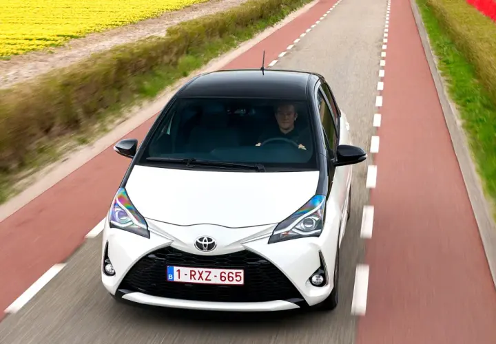 Biały Toyota Yaris Sedan z czarnym dachem jedzie drogą wśród pól żółtych kwiatów i czerwonych tulipanów.