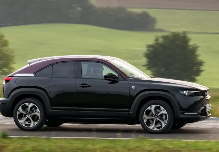 Czarna Mazda MX-30 z bordowym dachem sunie po drodze wśród zielonych wzgórz.
