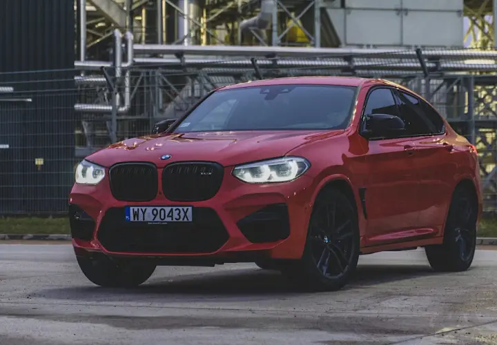 Czerwone BMW X4 z czarnymi felgami i grillem, zaparkowane przed przemysłowym budynkiem.