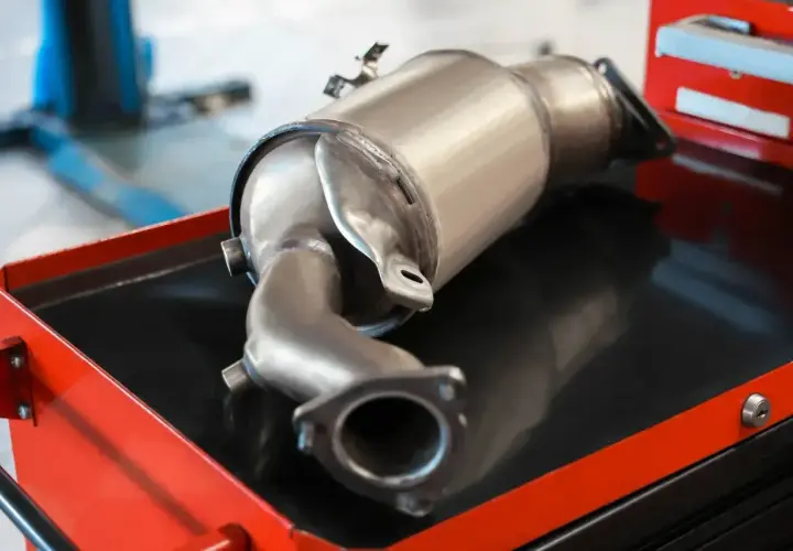 Metalowy element układu wydechowego samochodu, prawdopodobnie filtr DPF, leżący na wózku warsztatowym.