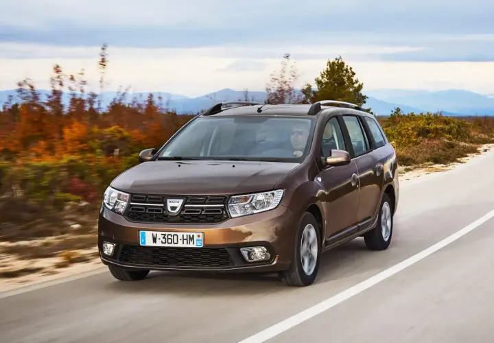 Brązowy Dacia Logan MCV jedzie po krętej drodze wśród jesiennych drzew i gór w tle.