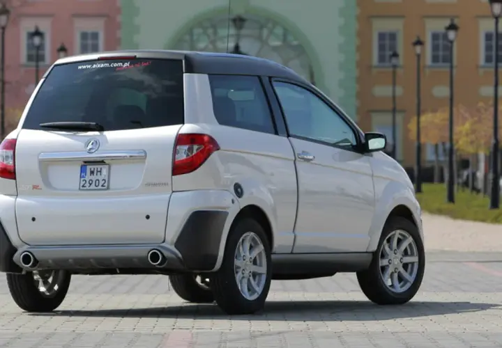 Białe auto Aixam GTR, idealne jako auto bez prawka, parkuje na brukowanym placu przed kolorowymi kamienicami.