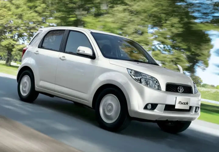 Biały Toyota Rush pędzi po drodze, otoczony zielenią drzew.