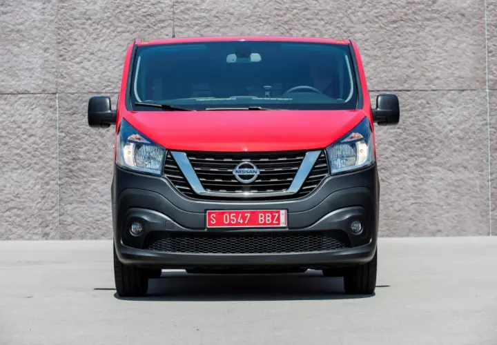 Czerwony Nissan NV300 z czarnym zderzakiem i chromowaną atrapą chłodnicy stoi przed betonową ścianą.