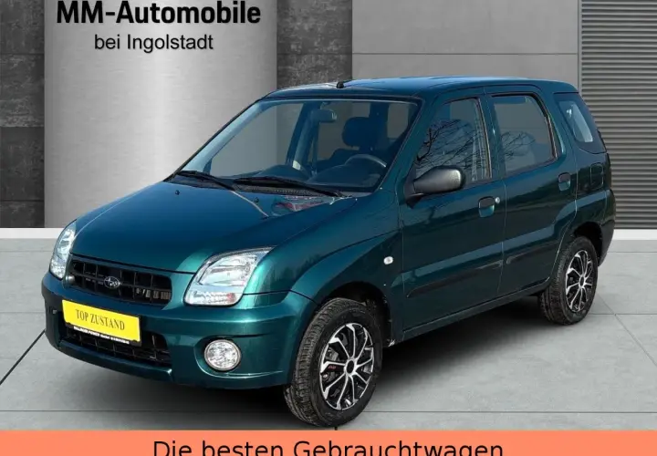 Zielony Subaru Justy z żółtą tabliczką "TOP ZUSTAND" na tle hali dealerskiej.