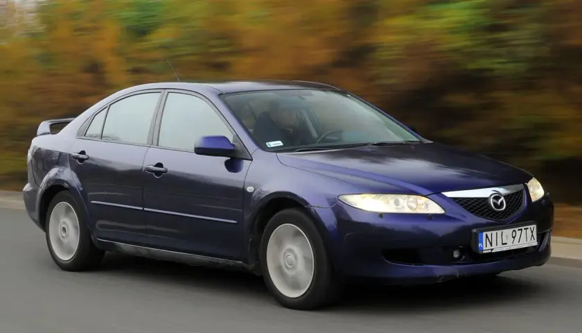 Granatowa Mazda 6 GG jedzie po drodze, za nią rozmyty krajobraz jesiennych drzew.