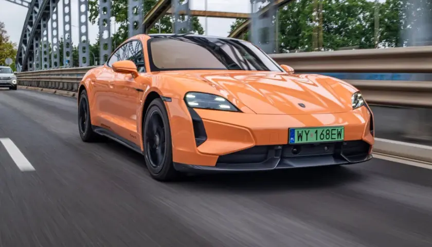 Pomarańczowy Porsche Taycan Turbo S pędzi po moście, jego sportowy design podkreślony przez dynamiczne linie.