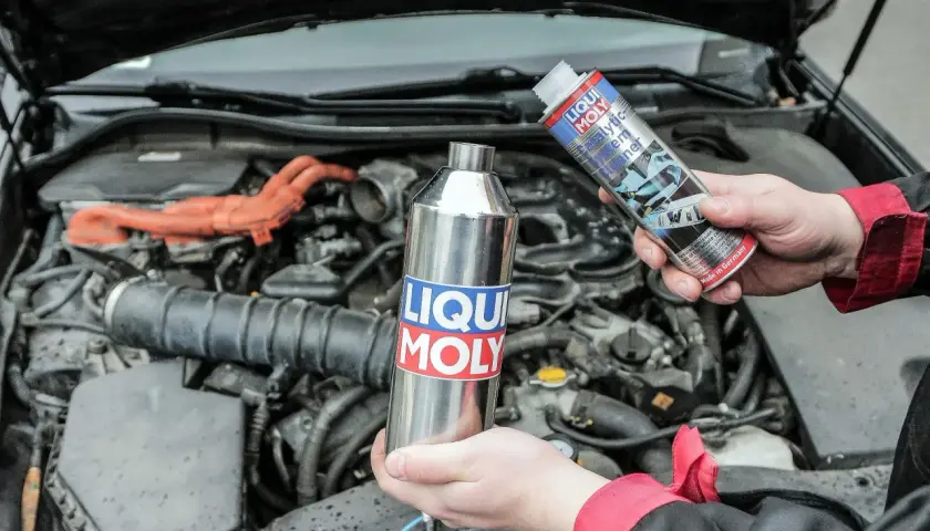Ręce trzymające preparaty Liqui Moly do czyszczenia katalizatora na tle silnika samochodu.