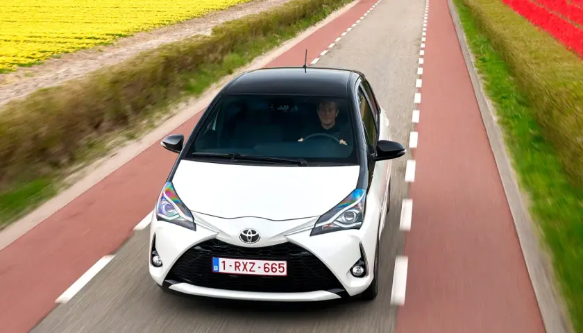 Biały Toyota Yaris Sedan z czarnym dachem jedzie drogą wśród pól żółtych kwiatów i czerwonych tulipanów.