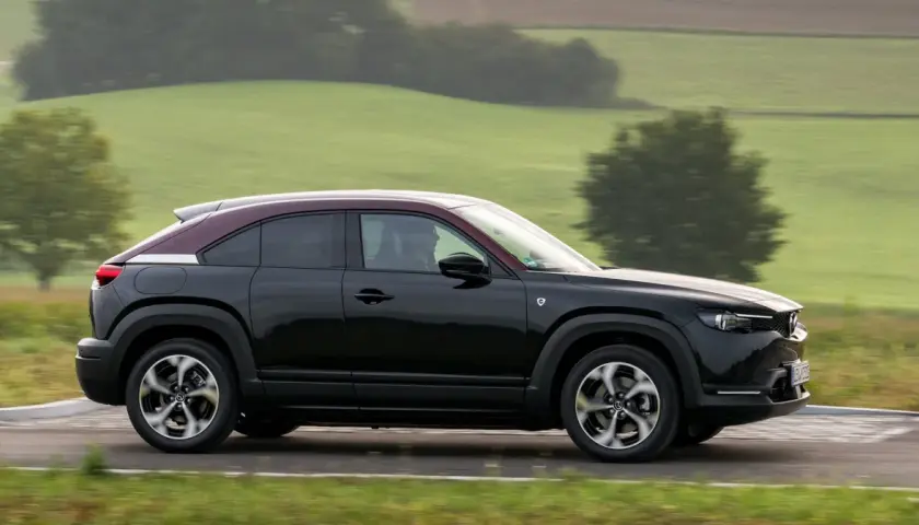 Czarna Mazda MX-30 z bordowym dachem sunie po drodze wśród zielonych wzgórz.