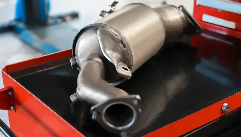 Metalowy element układu wydechowego samochodu, prawdopodobnie filtr DPF, leżący na wózku warsztatowym.