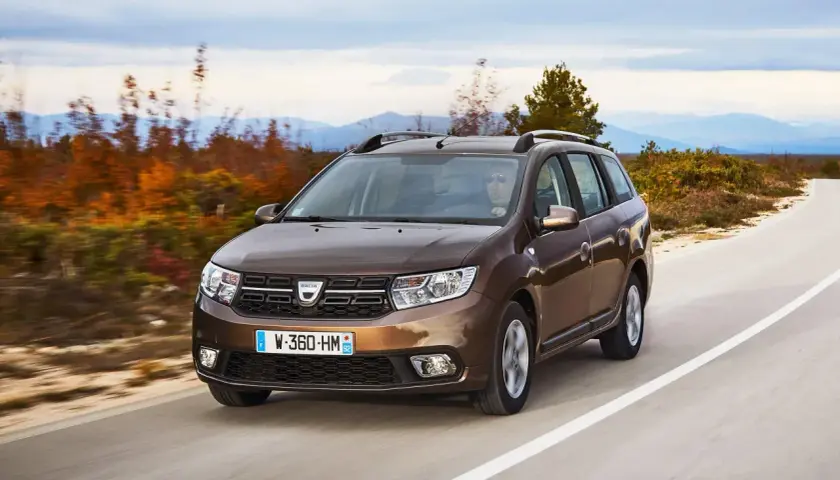Brązowy Dacia Logan MCV jedzie po krętej drodze wśród jesiennych drzew i gór w tle.