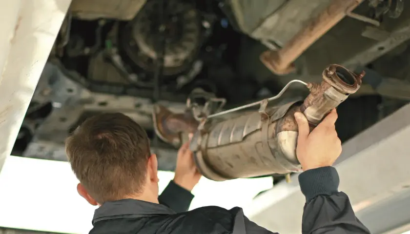 Mechanik trzymający pod samochodem zardzewiały element układu wydechowego, prawdopodobnie DPF.