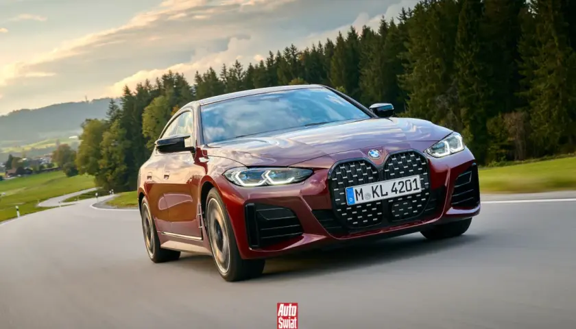 Czerwone BMW G26 z charakterystycznym grillem pędzi po krętej drodze wśród zielonych wzgórz i lasów.