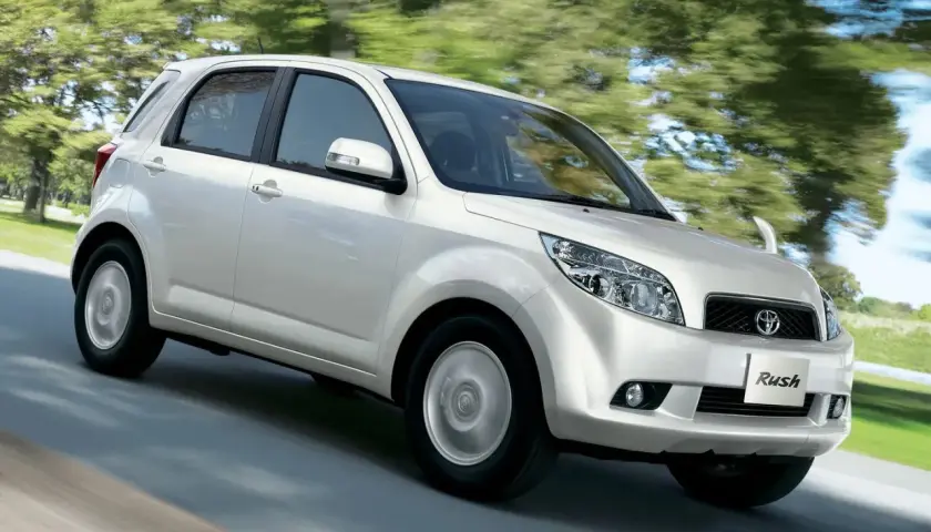 Biały Toyota Rush pędzi po drodze, otoczony zielenią drzew.