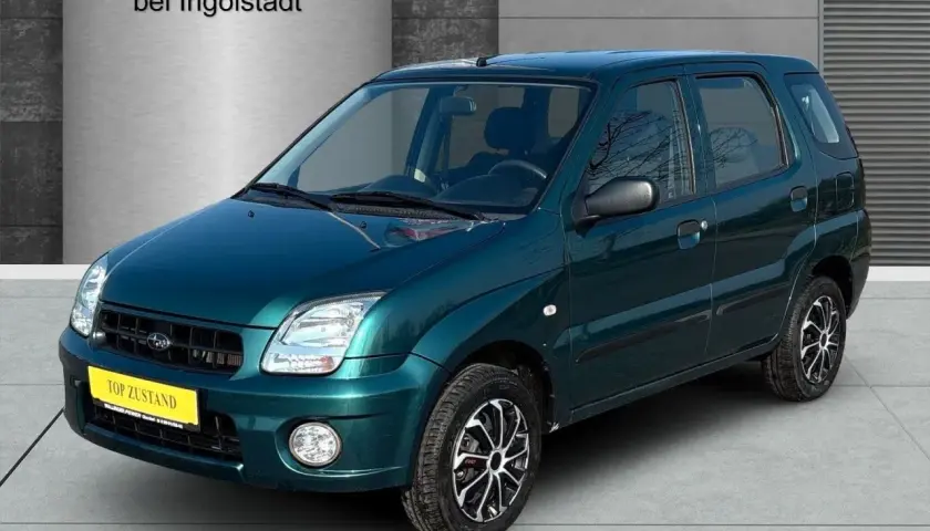 Zielony Subaru Justy z żółtą tabliczką "TOP ZUSTAND" na tle hali dealerskiej.