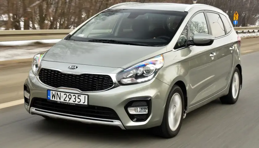 Srebrny Kia Carens 4 w ruchu drogowym, z widocznym grillem i reflektorami.