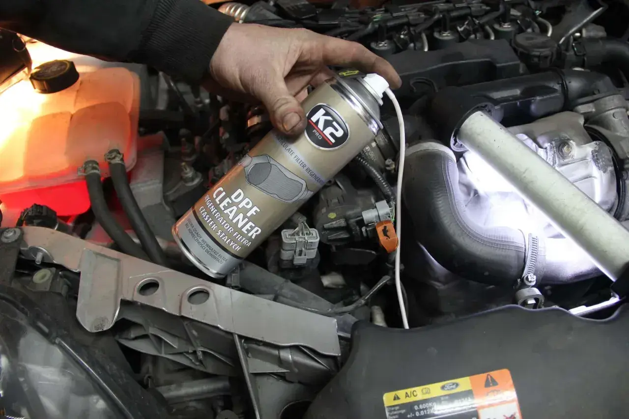Ręka trzymająca puszkę preparatu do czyszczenia DPF na tle silnika samochodowego.