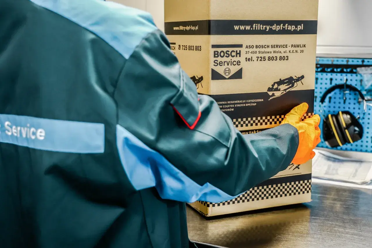 Pracownik w kombinezonie i rękawiczkach trzyma karton z logo Bosch Service.