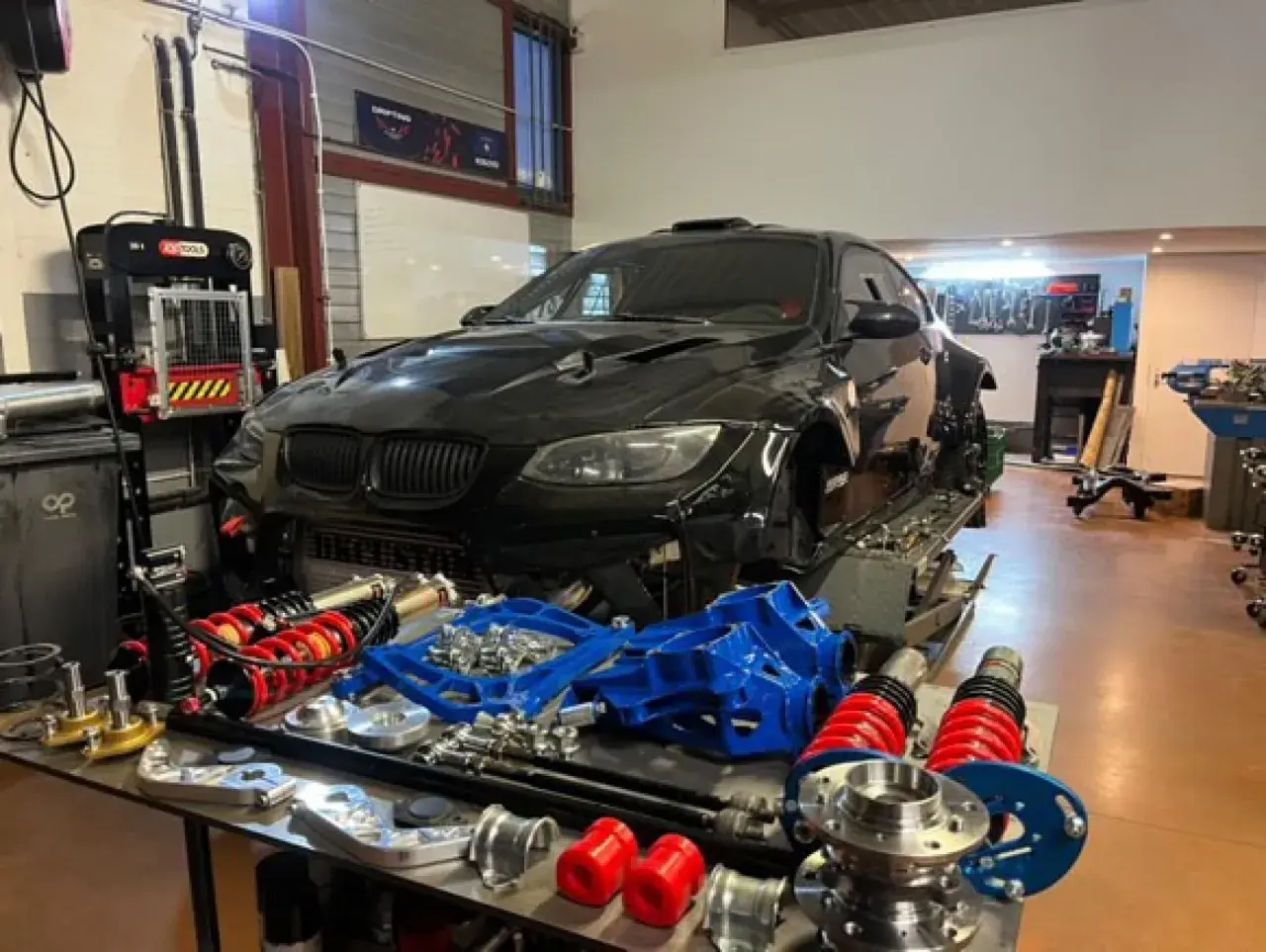 Warsztat z czarnym BMW E92, częściami zawieszenia i narzędziami. Lata produkcji tego modelu to 2006-2013.