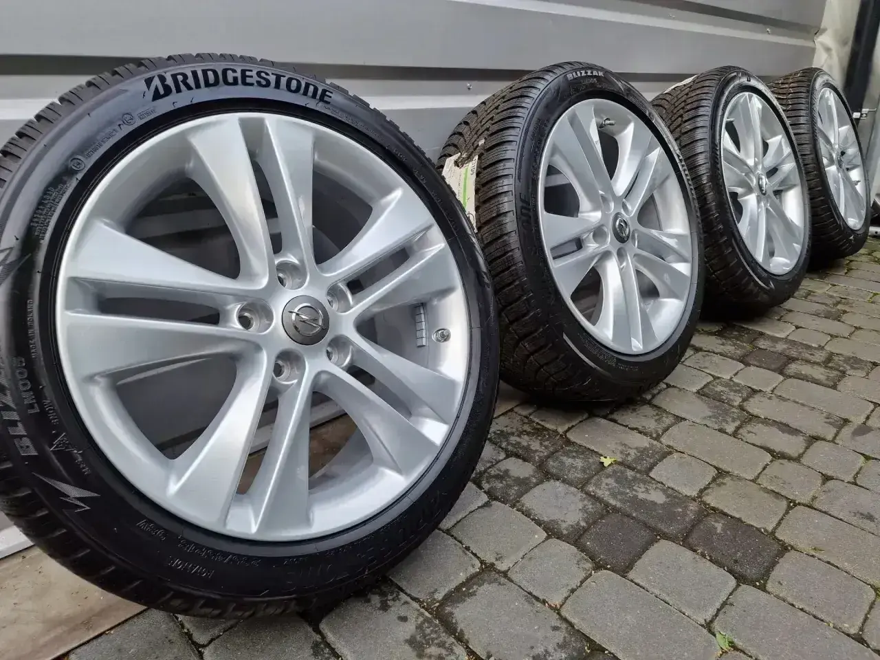 Cztery felgi aluminiowe z oponami zimowymi Bridgestone, z logo Opla, stojące na bruku.